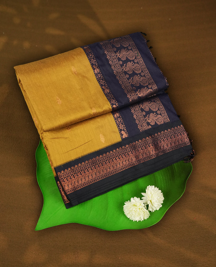 Mustard yellow colour Kalyani Cotton saree with subtle zari woven motifs all over, paired with a contrasting navy blue rich copper zari border & peacock motifs on pallu & Comes with a matching blouse piece.