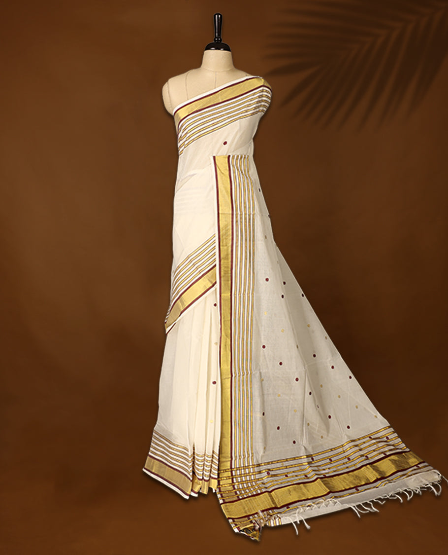 Off white colour pure handloom soft cotton kerala saree with maroon and mustard polka motifs, highlighted by a golden zari border and striped pallu in gold, maroon, and green, and paired with a matching blouse, perfect for festivities.