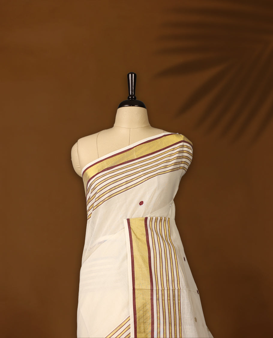 Off white colour pure handloom soft cotton kerala saree with maroon and mustard polka motifs, highlighted by a golden zari border and striped pallu in gold, maroon, and green, and paired with a matching blouse, perfect for festivities.