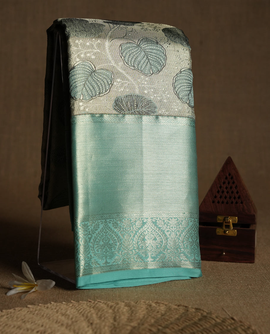 Silver Colour Kancheepuram tissue silk saree featuring intricate silver and blue zari leaf motifs on the body, gracefully complemented by a light blue traditional peacock and floral design border, and paired with a matching blouse.