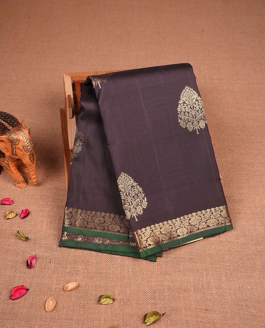 Deep coffee brown colour pure traditional Kancheepuram silk saree adorned with elegant zari tree motifs across the body, complemented by a rich zari woven border with a subtle green edge, paired with a matching blouse piece.