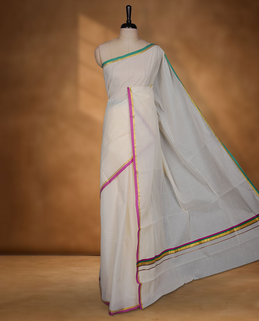 Off-white colour cotton Kerala saree, featuring a classic gold kasavu border highlighted with subtle green and magenta accents, finished with a soft, airy drape and paired with a matching blouse.