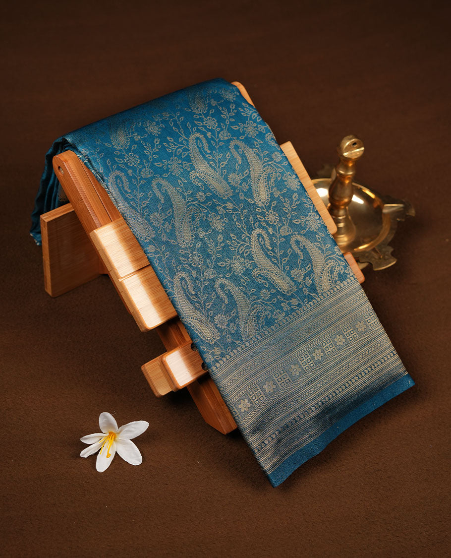 Peacock blue colour semi soft silk brocade saree featuring elegant gold zari floral and paisley motifs across the body, finished with a richly woven self-colour border and pallu, paired with a matching blouse.