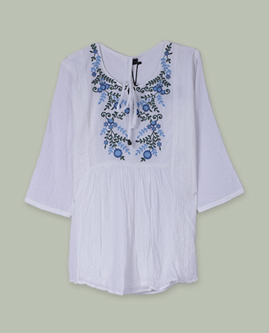 White three-quarter sleeve tunic top with round neckline, front yoke featuring blue and green floral embroidery, and tie-up detail at the neck.