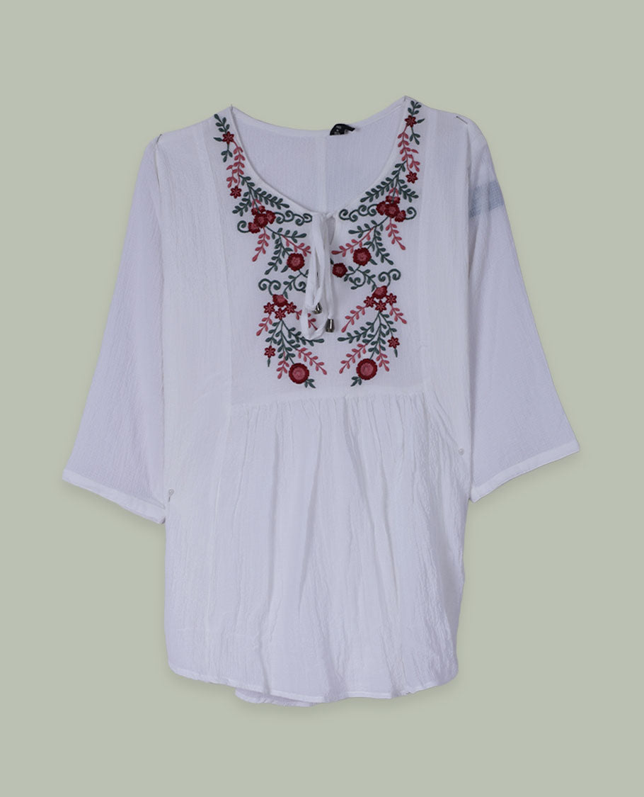 White three-quarter sleeve tunic top with round neckline and front yoke featuring red and green floral embroidery with tie-up detail at the neck.