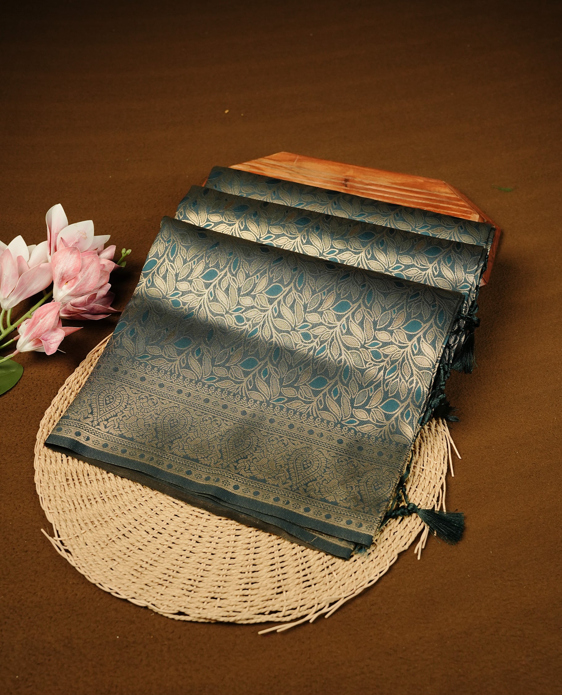 Teal Blue Colour Softie Silk Saree Featuring An All Over Gold Woven Leaf Pattern, Finished With A Matching Border Adorned With Intricate Paisley And Floral Motifs, Paired With A Matching Blouse Piece.