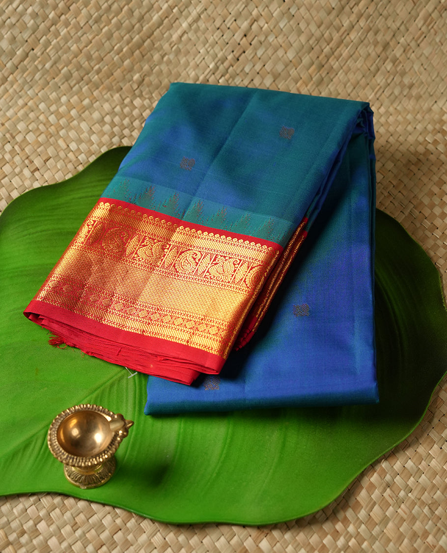 Peacock colour Vasundhara silk saree adorned with all-over gold mayil motifs on the body, complemented by a contrast red colour with gold traditional with mayil pattern border and paired with a matching blouse piece.