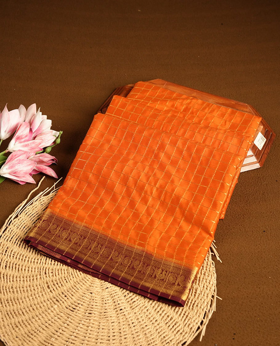 Orange colour mysore crepe saree with gold zari check pattern on body, complemented by contrast Chocolate brown colour border featuring peacock and chakkram motifs, paired with matching blouse.