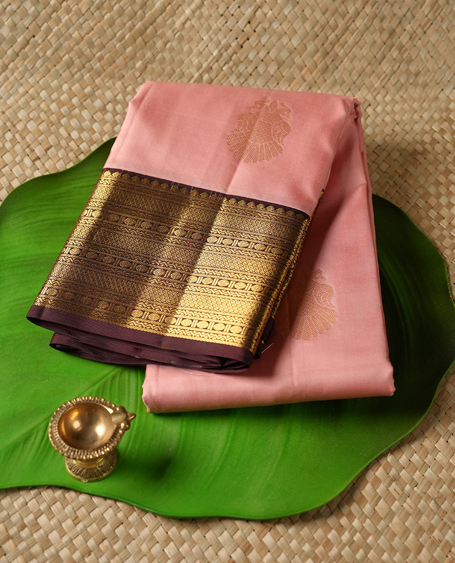 Baby pink colour Vasundhara silk saree adorned with all-over gold two head mayil motifs on the body, complemented by a contrast brown colour with gold traditional border and paired with a matching blouse piece.