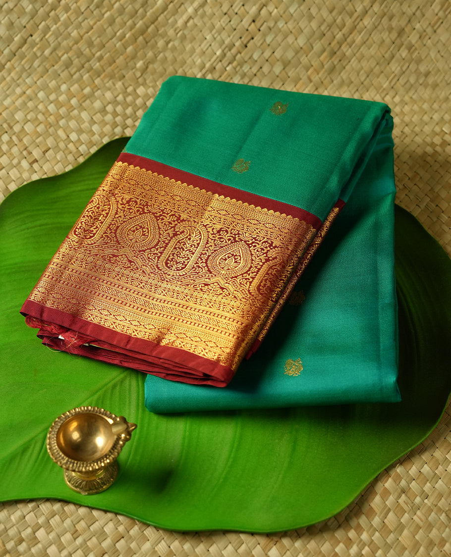 Green colour Vasundhara silk saree adorned with all-over gold mayil motifs on the body, complemented by a contrast maroon colour with gold traditional border and paired with a matching blouse piece.