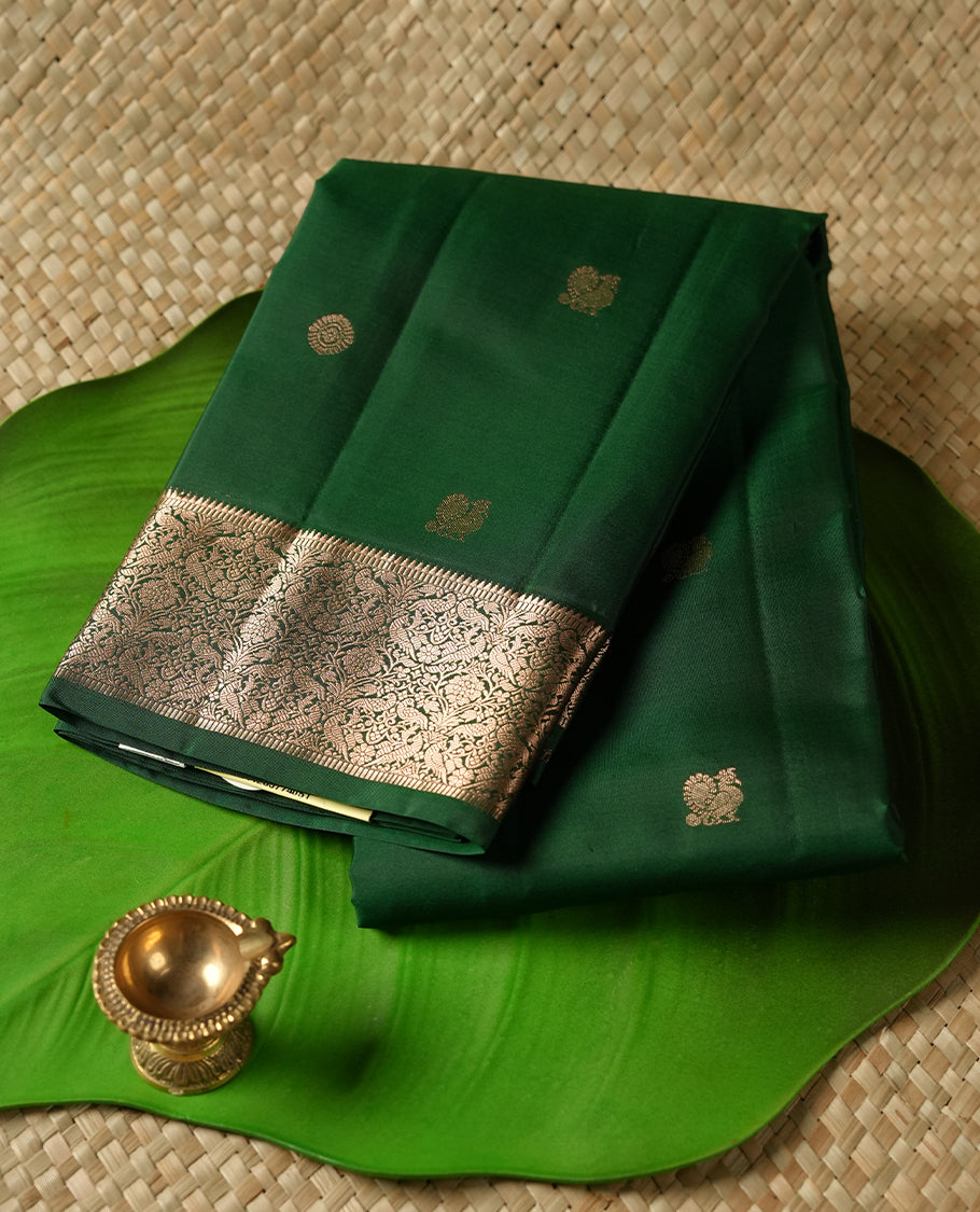 Dark Green colour Vasundhara silk saree adorned with all-over gold chakkaram & mayil motifs on the body, complemented by a gold tarditional border and paired with a matching blouse piece.