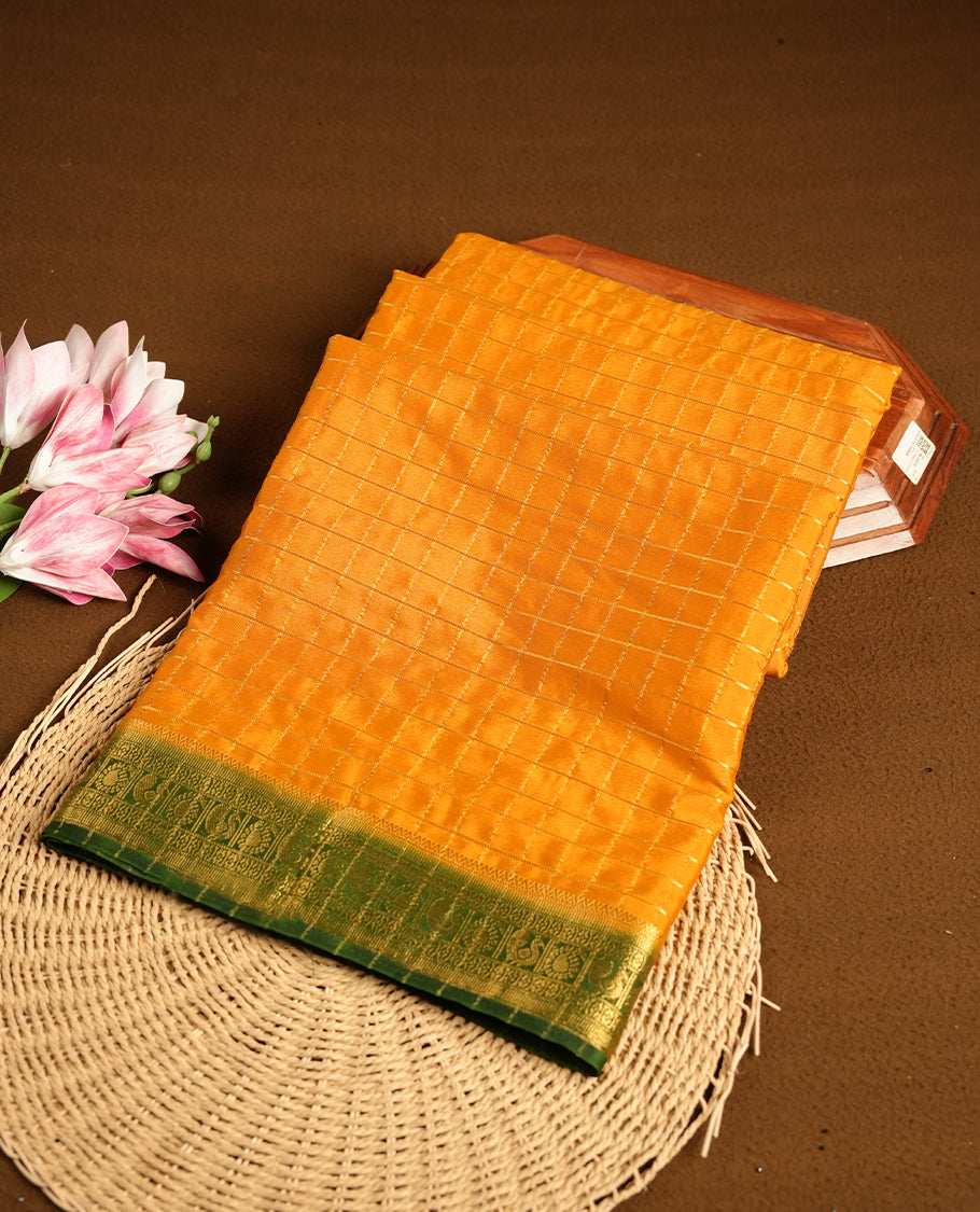 Mustard colour mysore crepe saree with gold zari check pattern on body, complemented by contrast Green colour border featuring peacock and chakkram motifs, paired with matching blouse.