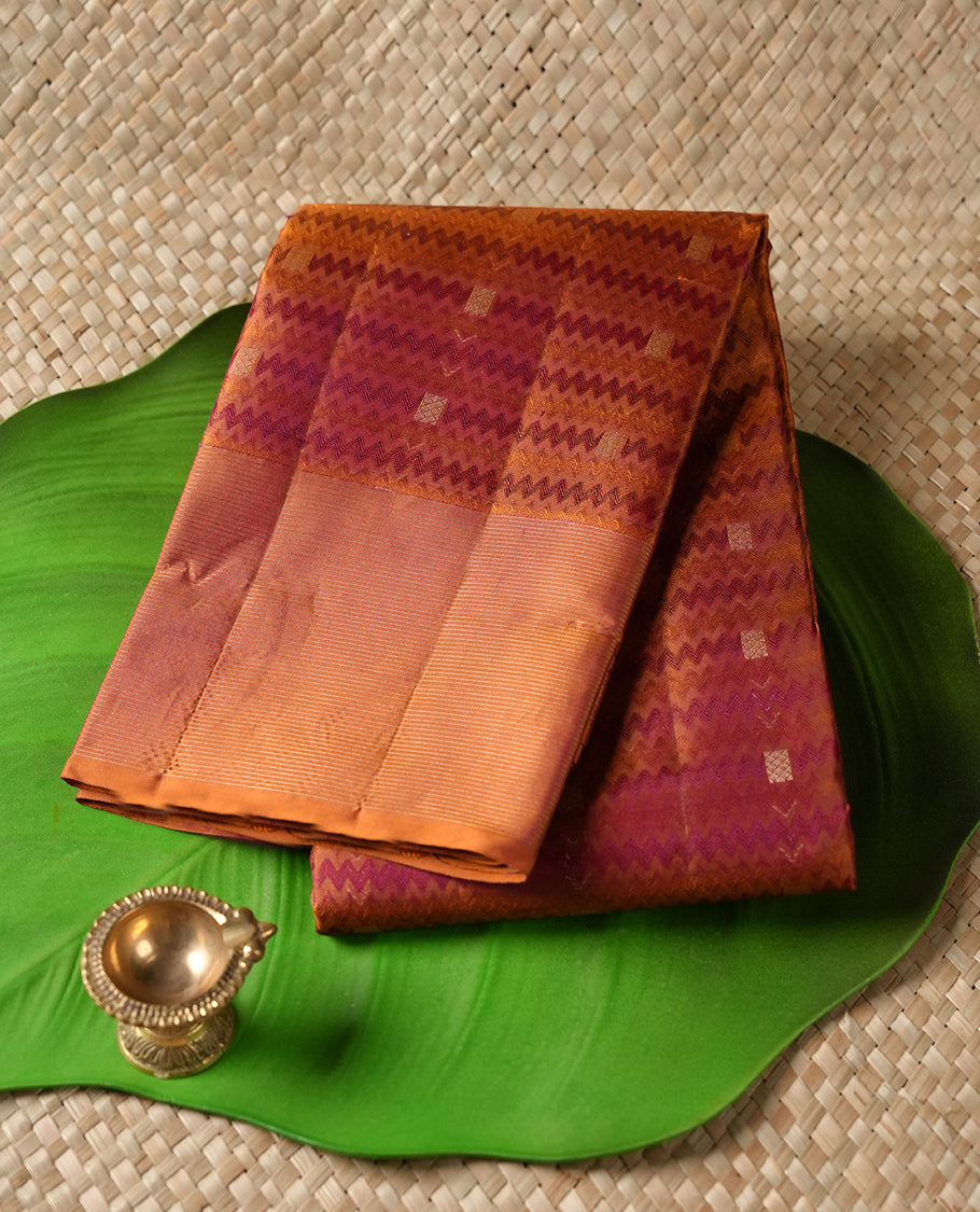 Burnt orange colour Vasundhara silk saree adorned with all-over purple zigzag motifs on the body, complemented by a self colour with gold line pattern border and paired with a matching blouse piece.