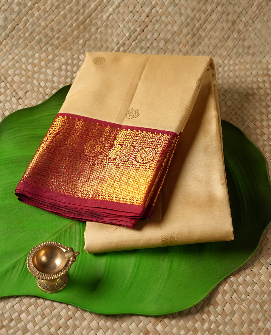 Cream colour Vasundhara silk saree adorned with all-over gold chakkaram & mayil motifs on the body, complemented by a contrast maroon gold tarditional border and paired with a matching blouse piece.