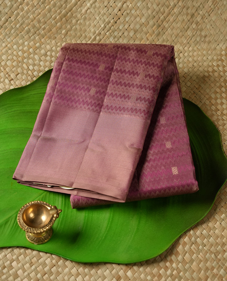 Dusty purple colour Vasundhara silk saree adorned with all-over zigzag motifs on the body, complemented by a self colour with gold line pattern border and paired with a matching blouse piece.
