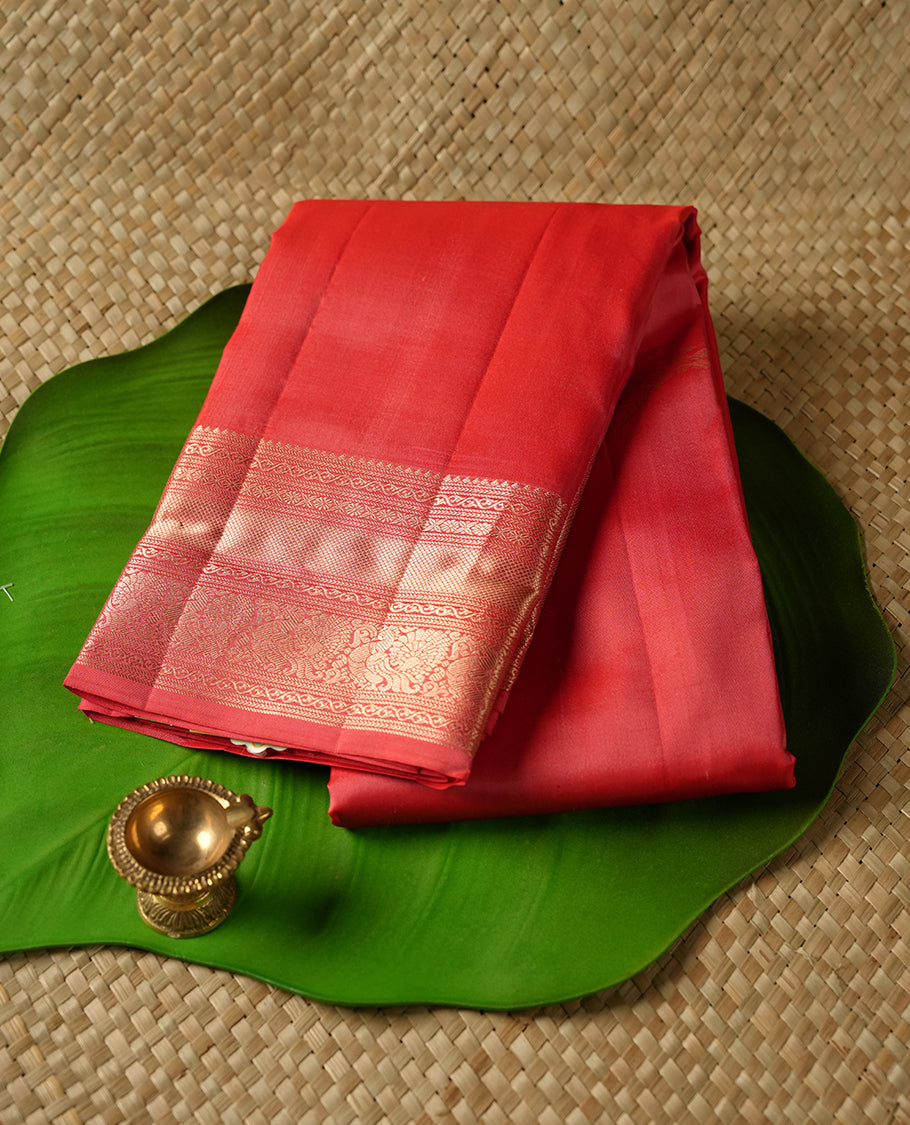 Reddish peach colour Vasundhara silk saree featuring Gold zari motifs across the body, finished with a self‑coloured traditional border and a coordinating blouse.