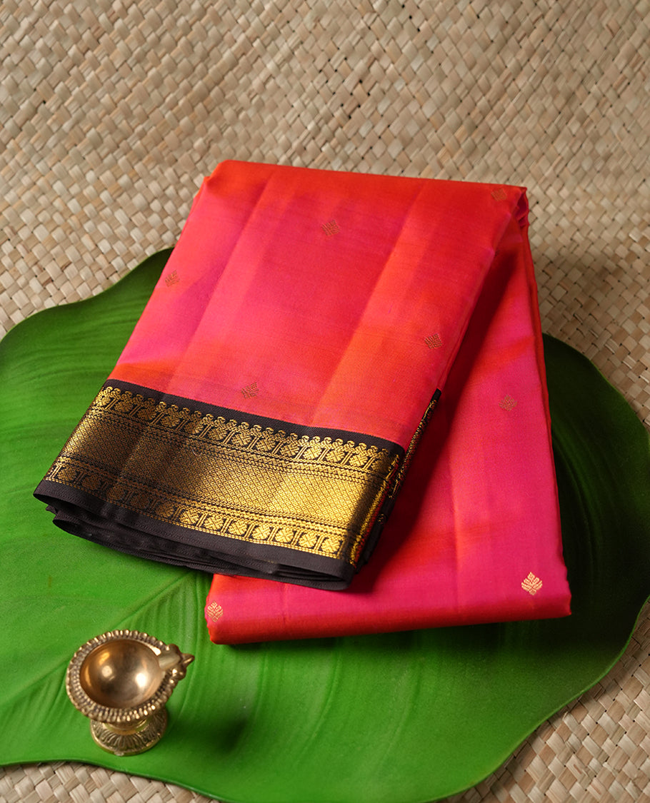 Pinkish red colour Vasundhara silk saree adorned with all-over gold zari motifs on the body, complemented by a contast black colour with gold motifs on border and paired with a matching blouse piece.