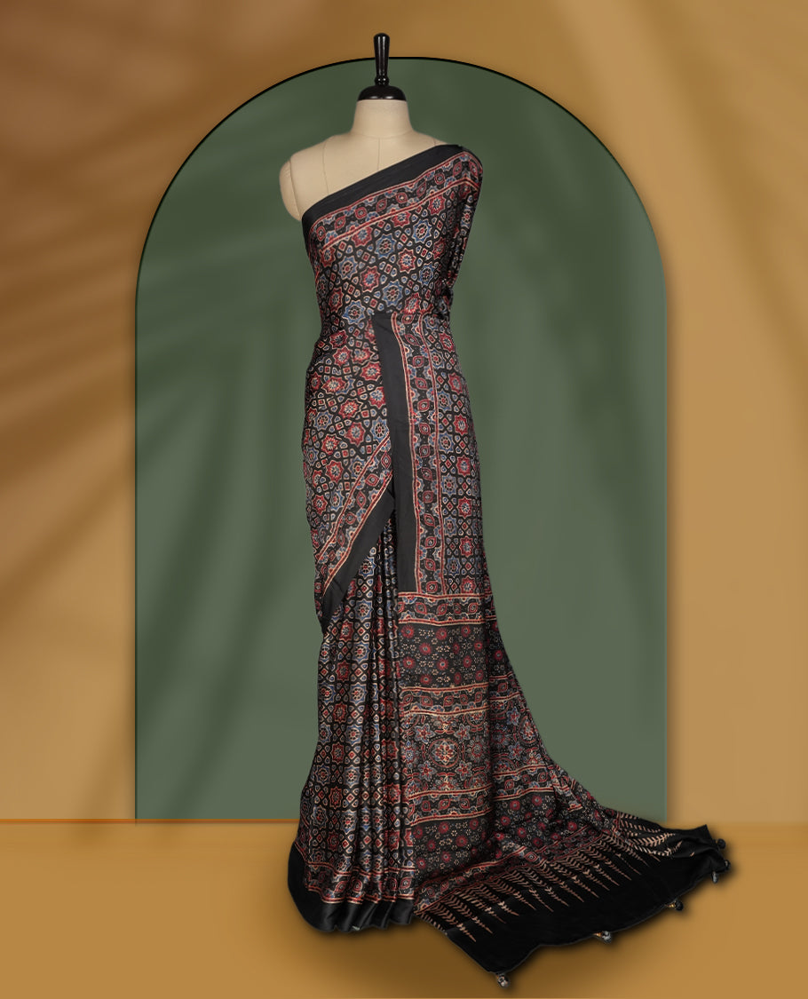 Black colour saree featuring intricate traditional prints in earthy tones, finished with a sleek satin border and tassel details & Comes with a stylish matching blouse.