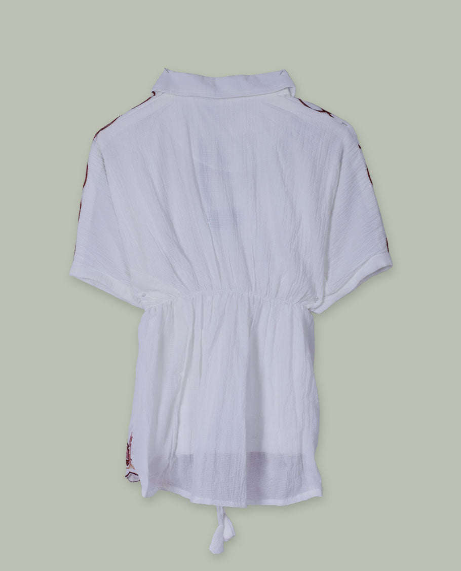 White short tunic top with collared neckline, short sleeves, contrast paisley and floral embroidery on shoulders and hem, and a drawstring waist with tassel ties.