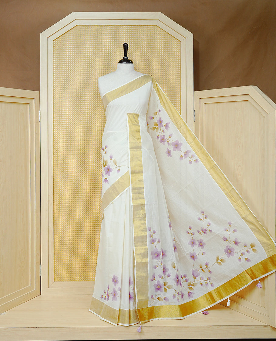 Cream Colour Cotton Kerala Saree Featuring Digital Purple Floral Prints Across The Body, Enhanced With A Gold Zari Border, Traditional Gold Zari Pallu, Finished With Purple & White Tassels And A Matching Blouse Piece