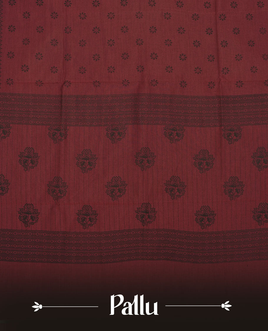 Deep maroon colour poly cotton saree with black floral butti motifs on the body, paired with a matching pallu featuring black ornate floral jaal and stripes on the border, and a coordinated all‑over black paisley printed blouse piece.