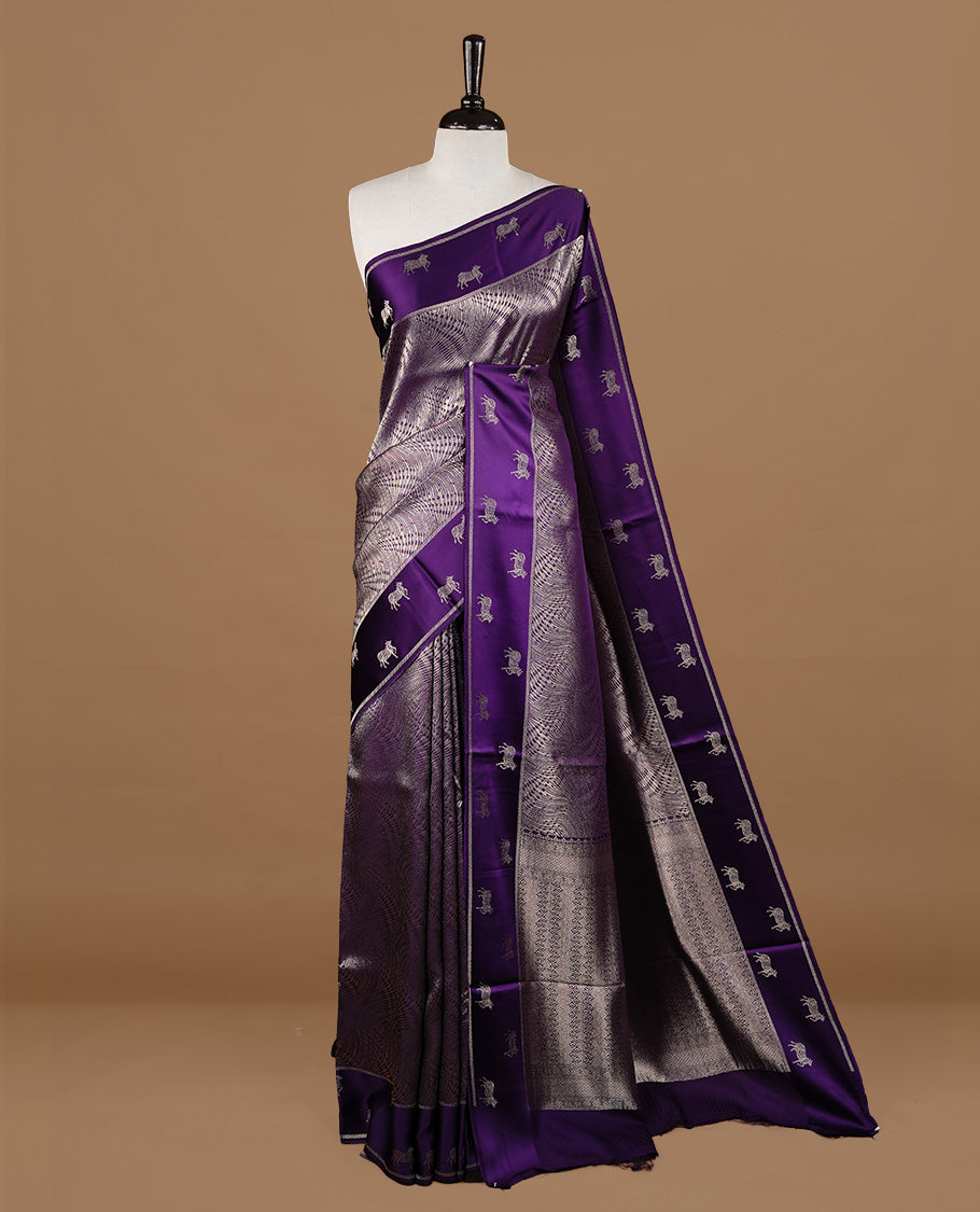 Purple colour semi Banaras crepe silk saree featuring an elegant gold zari patterned body, highlighted by a plain border adorned with traditional gold zari cow motifs. Paired with a matching blouse piece.