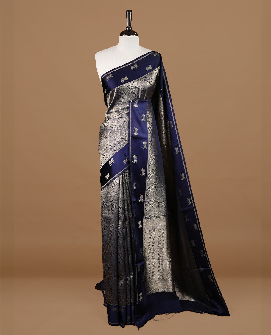 Dark Blue colour semi Banaras crepe silk saree featuring an elegant gold zari patterned body, highlighted by a plain border adorned with traditional gold zari cow motifs. Paired with a matching blouse piece.