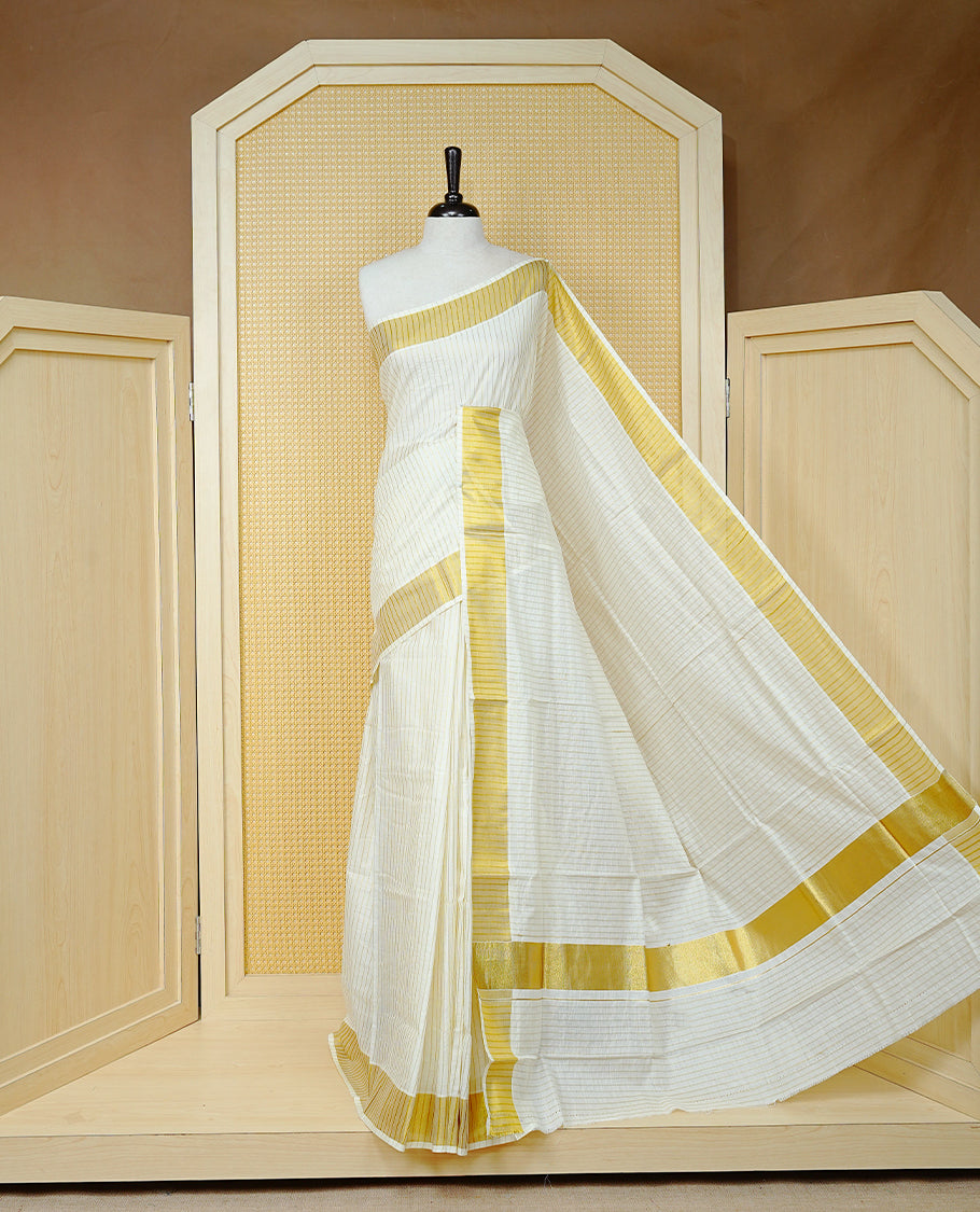 Cream Colur Cotton Kerala Saree Featuring Gold Zari Strips Across On The Body, Classic Gold Zari Border & Pallu,Paired With A Matching Blouse Piece.