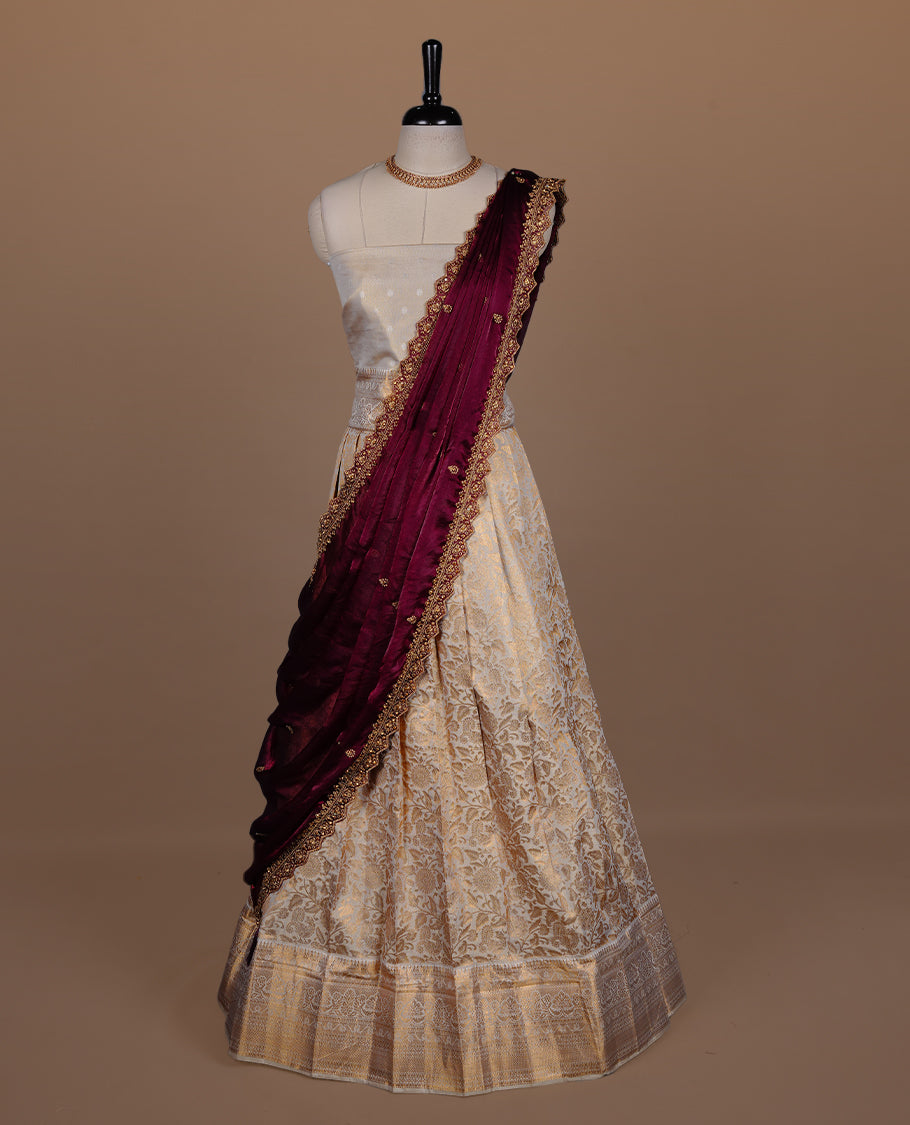 Light gold colour semi banaras semi stitched dhavani set featuring gold zari floral kodi motifs and traditional border, unstitched matching blouse piece and contrast Maroon colour Duptta with gold thread and beats worked border.