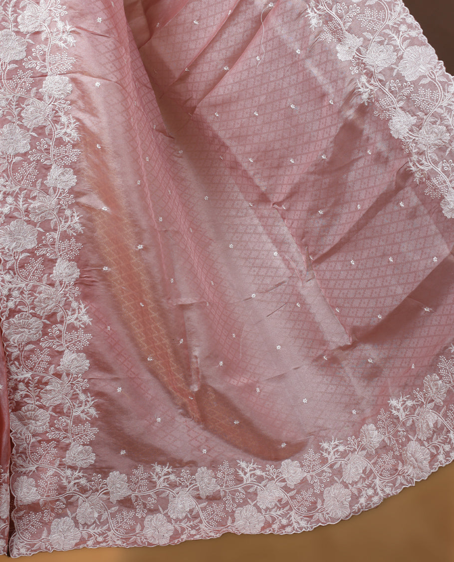 Pastel peach colour organza designer saree featuring a subtle self patterned body and an intricately scalloped floral thread embroidered border all around, complemented by a coordinating embroidered pallu and paired with a matching blouse.