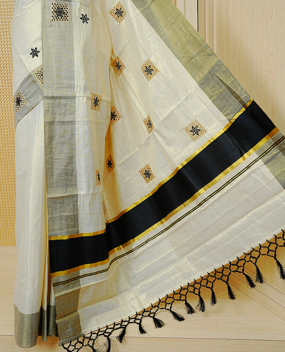 Cream Colour Tissue Kerala Saree Featuring Delicate Black Floral Embroidery Work Across The Body, Enhanced With A Grey Zari Border And A Rich Black & Gold Zari Pallu, Finished With Black Tassels And A Matching Blouse Piece