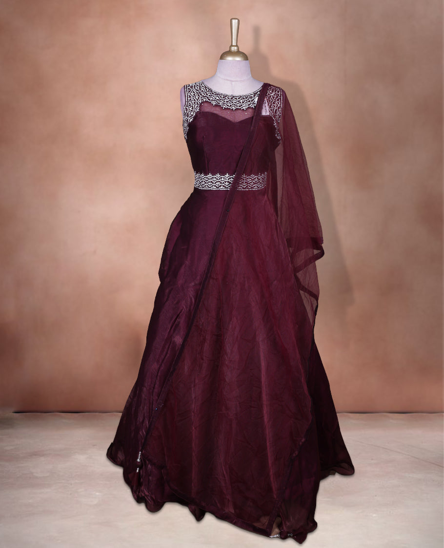 Wine colour Gown with stitched sleeveless embellished blouse featuring sheer yoke and waist detailing, matching stitched flared lehenga, and plain sheer dupatta.