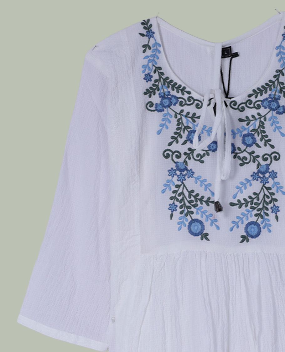 White three-quarter sleeve tunic top with round neckline, front yoke featuring blue and green floral embroidery, and tie-up detail at the neck.