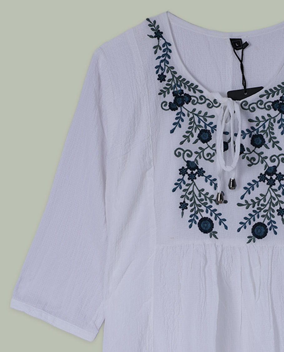 White three-quarter sleeve tunic top with round neckline and front yoke featuring teal and green floral embroidery with tie-up detail at the neck.
