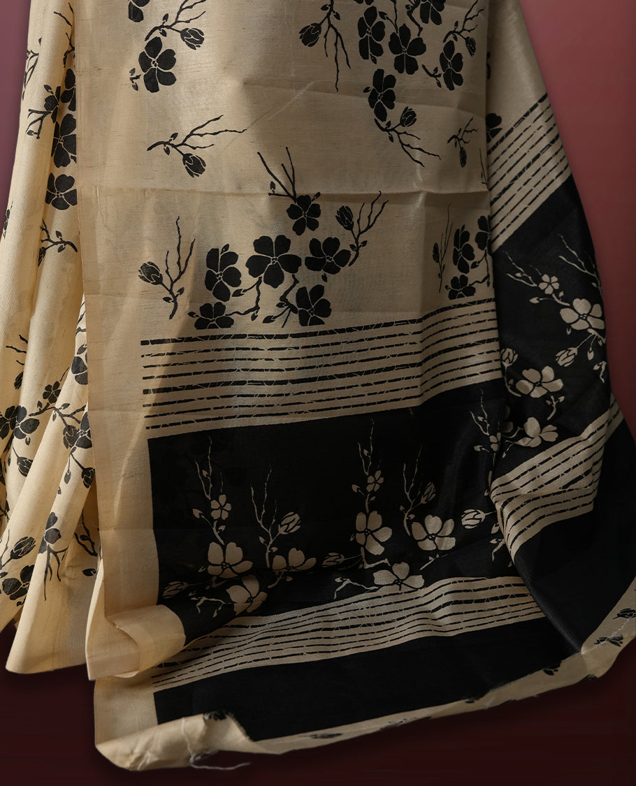 Beige colour saree adorned with black floral prints across the body, complemented by a striking black pallu with floral and stripe detailing. Finished with a subtle border and paired with a matching blouse.