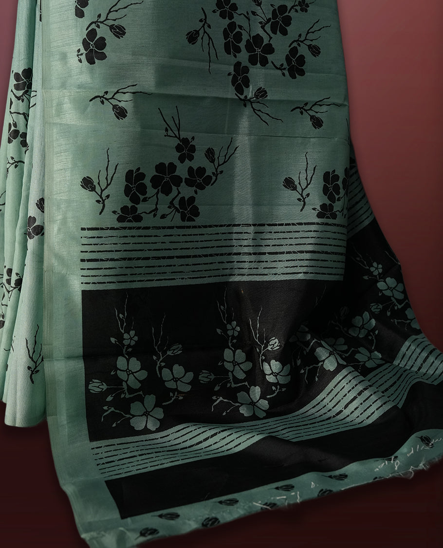 Pastel green colour saree with black floral prints all over the body, complemented by a contrasting black pallu adorned with bold floral designs and striped detailing & Paired with a matching blouse for a complete look.