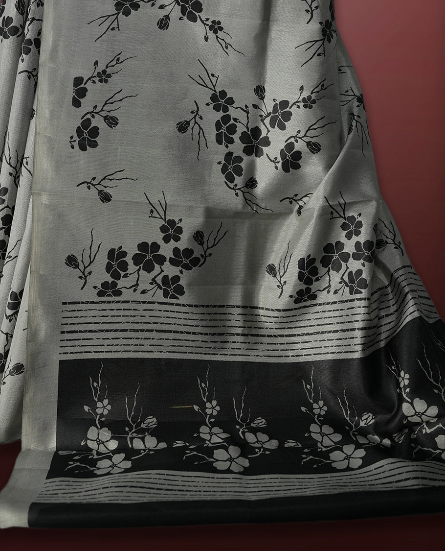 Grey colour saree with black floral prints all over the body, complemented by a contrasting black pallu adorned with bold floral designs and striped detailing & Paired with a matching blouse for a complete look.
