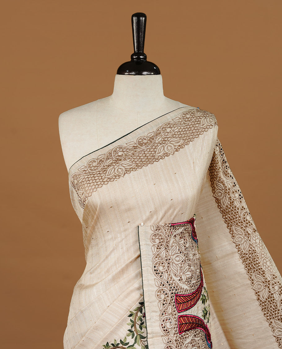 Cream colour Semi Tussar silk saree features an intricate cutwork embroidered peacock motifs on border, paired with a matching unstitched blouse piece.