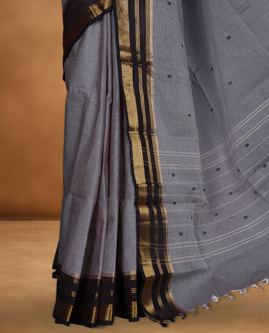 Grey colour chettinad cotton saree featuring small black buttas on body highlighting contrast dark chocolate colour with gold zari traditional border, pallu and paired with matching blouse piece.