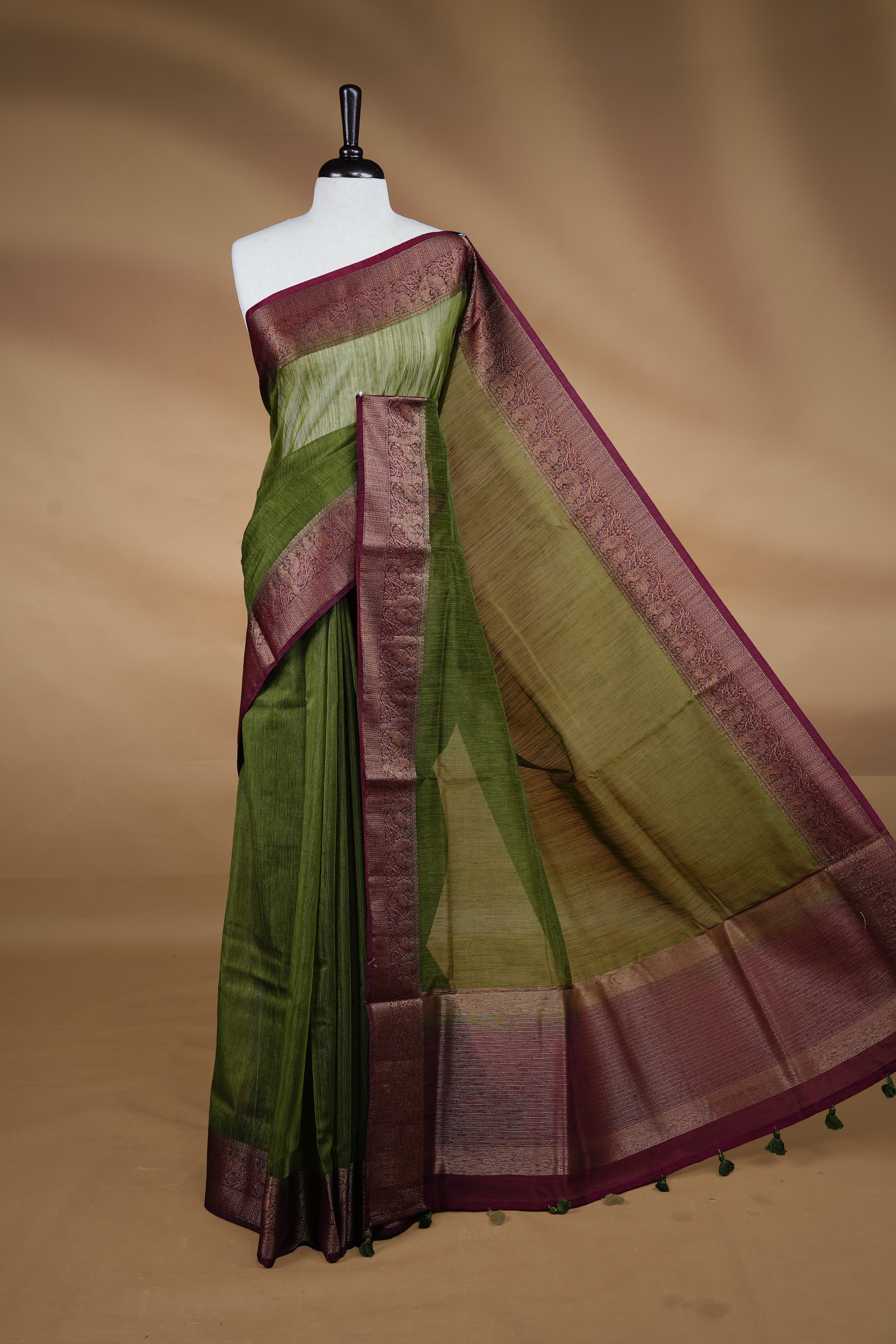 Olive green colour Semi linen saree featuring Elegant plain body, complemented by contrast Maroon colour With Antique zari border, paired with matching blouse piece.