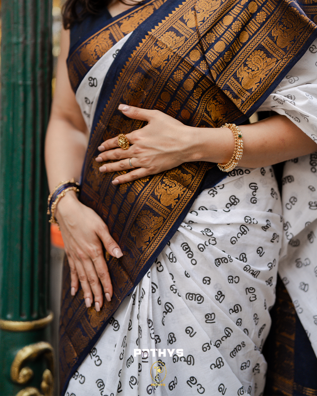 Off White Sungudi Cotton Saree featuring Malayalam alphabets on the body, a navy blue border with gold zari peacock and chakra motifs, a contrast pallu, and a matching blouse piece