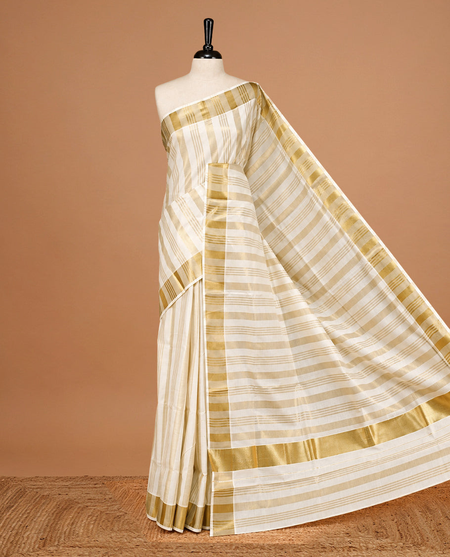 Cream Colour Cotton Kerala Saree Featuring Gold zari stripes On The Body & Classy Gold zari border, Paired With A Matching Blouse Piece.