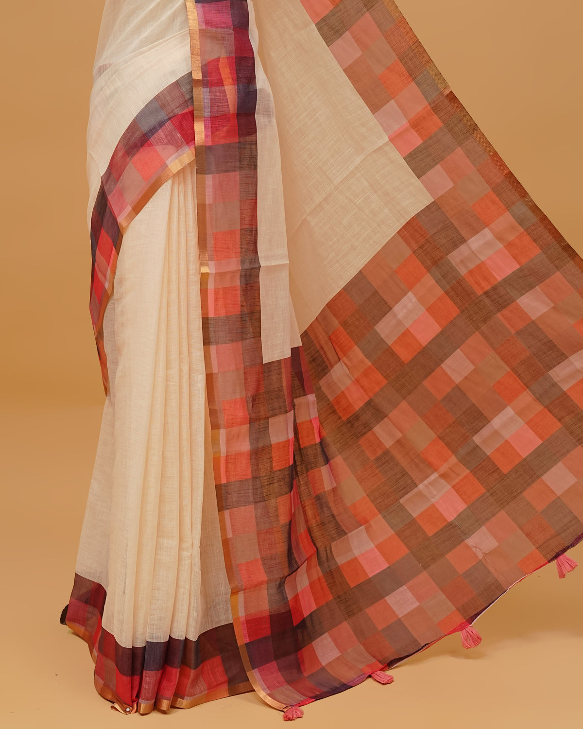 Cream colour semi linen saree featuring a plain body, a multicoloured check design with a gold tissue border, and a pallu finished with tassels. Paired with a matching blouse piece.