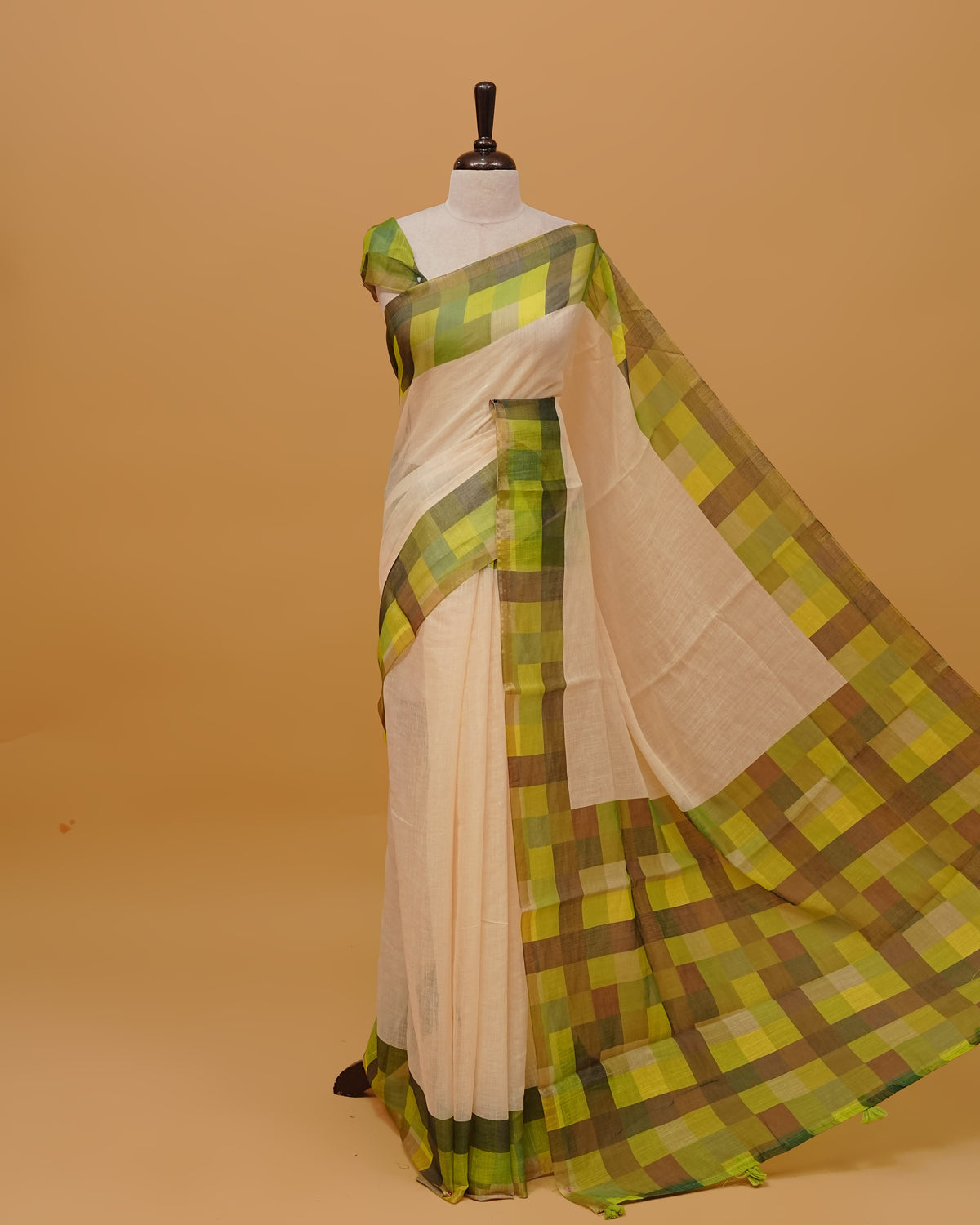 Cream colour semi linen saree featuring a plain body, a multicoloured check design with a gold tissue border, and a pallu finished with Tassels. Paired with a matching blouse piece.