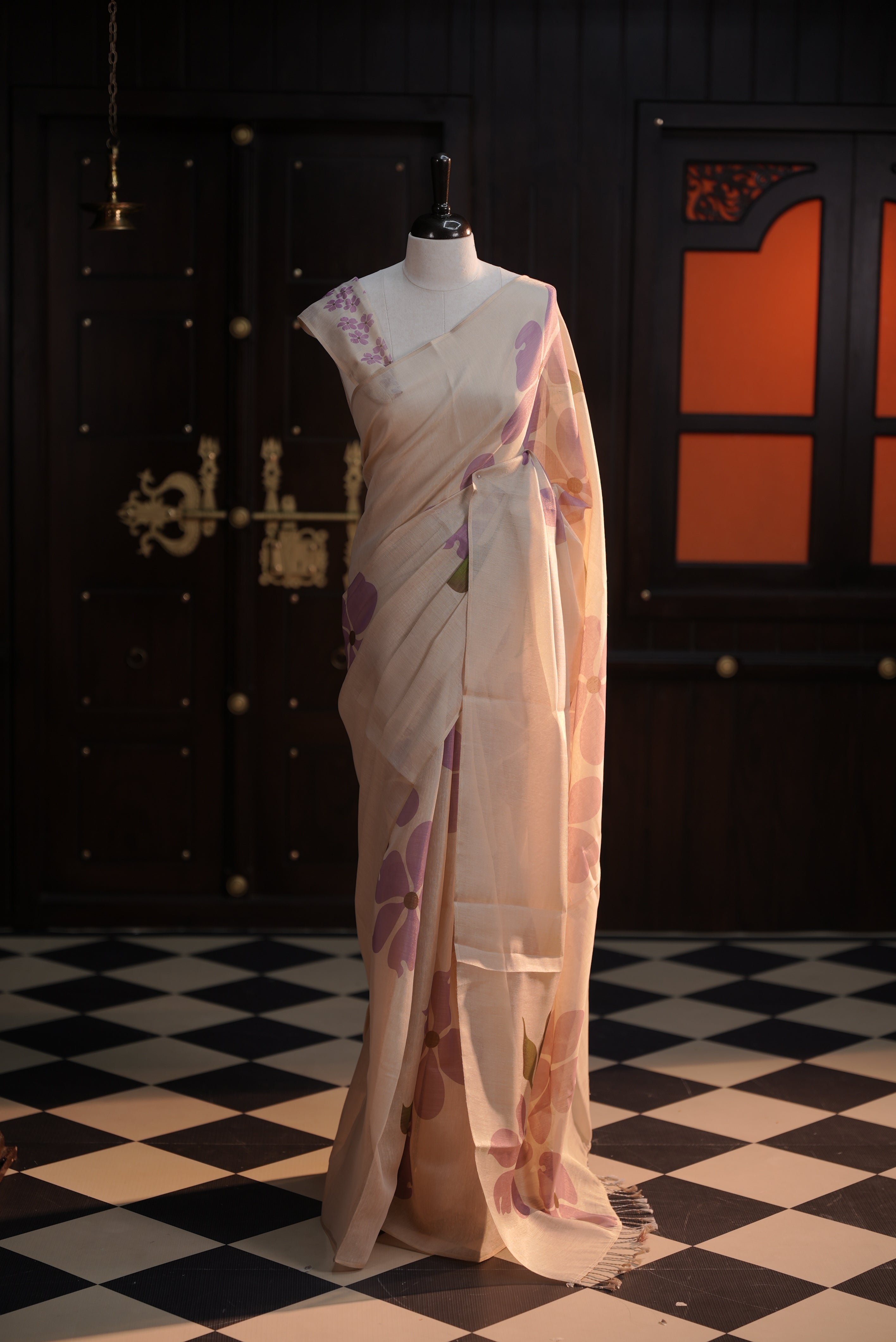 Beige colour semi soft tissue saree adorned with bold Lavender floral prints across the body, designed without a border, paired with a matching blouse piece featuring delicate flower motifs.