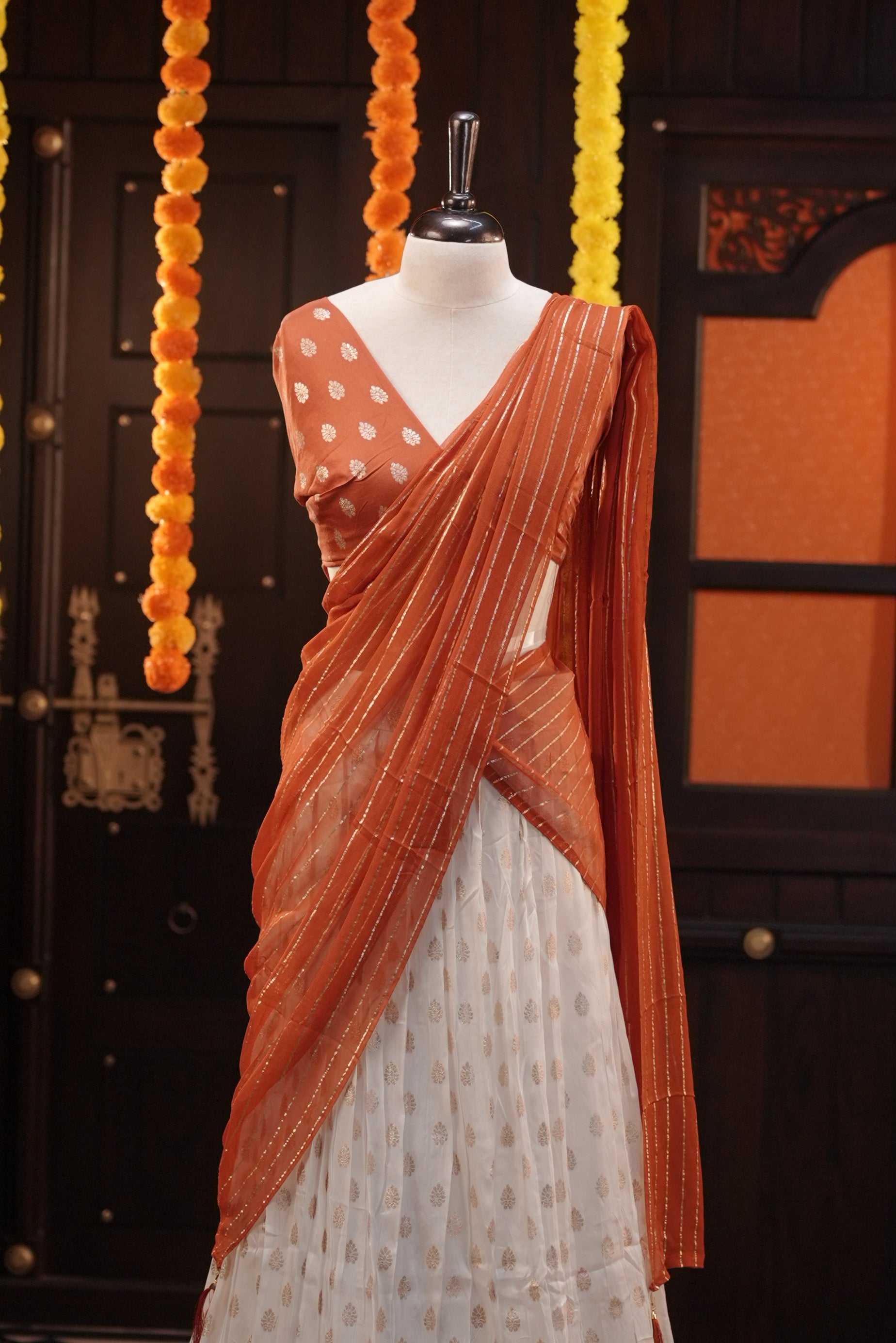 Off white colour Semi Stitched Banaras Dhavani Set Featuring off white Skirt with Gold zari buttas & Traditional border, paired with Orange colour Blouse material with gold zari buttas, finishing with same colour with gold zari Stripped Dupatta.