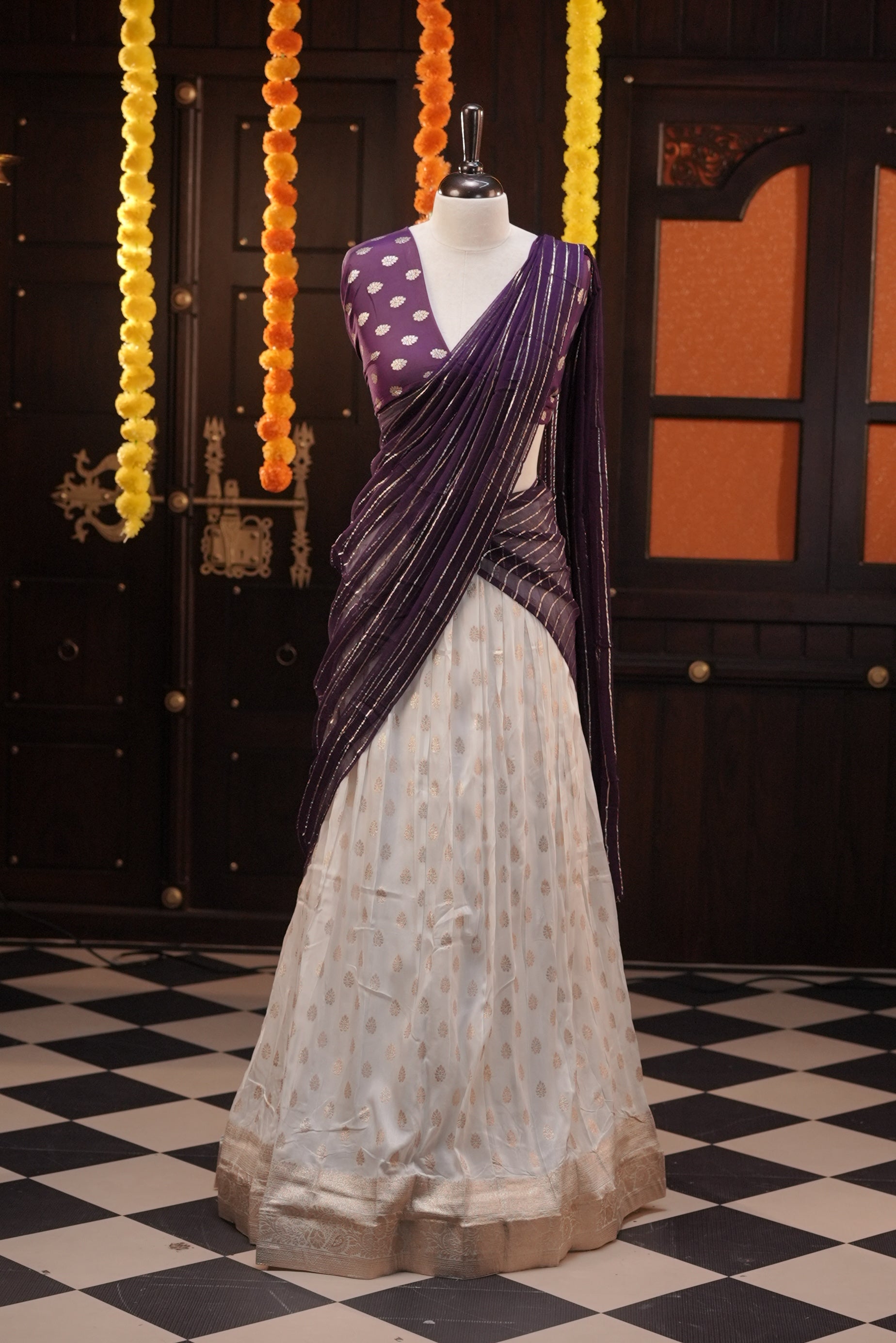 Off white colour Semi Stitched Banaras Dhavani Set Featuring off white Skirt with Gold zari buttas & Traditional border, paired with Purple colour Blouse material with gold zari buttas, finishing with same colour with gold zari Stripped Dupatta.