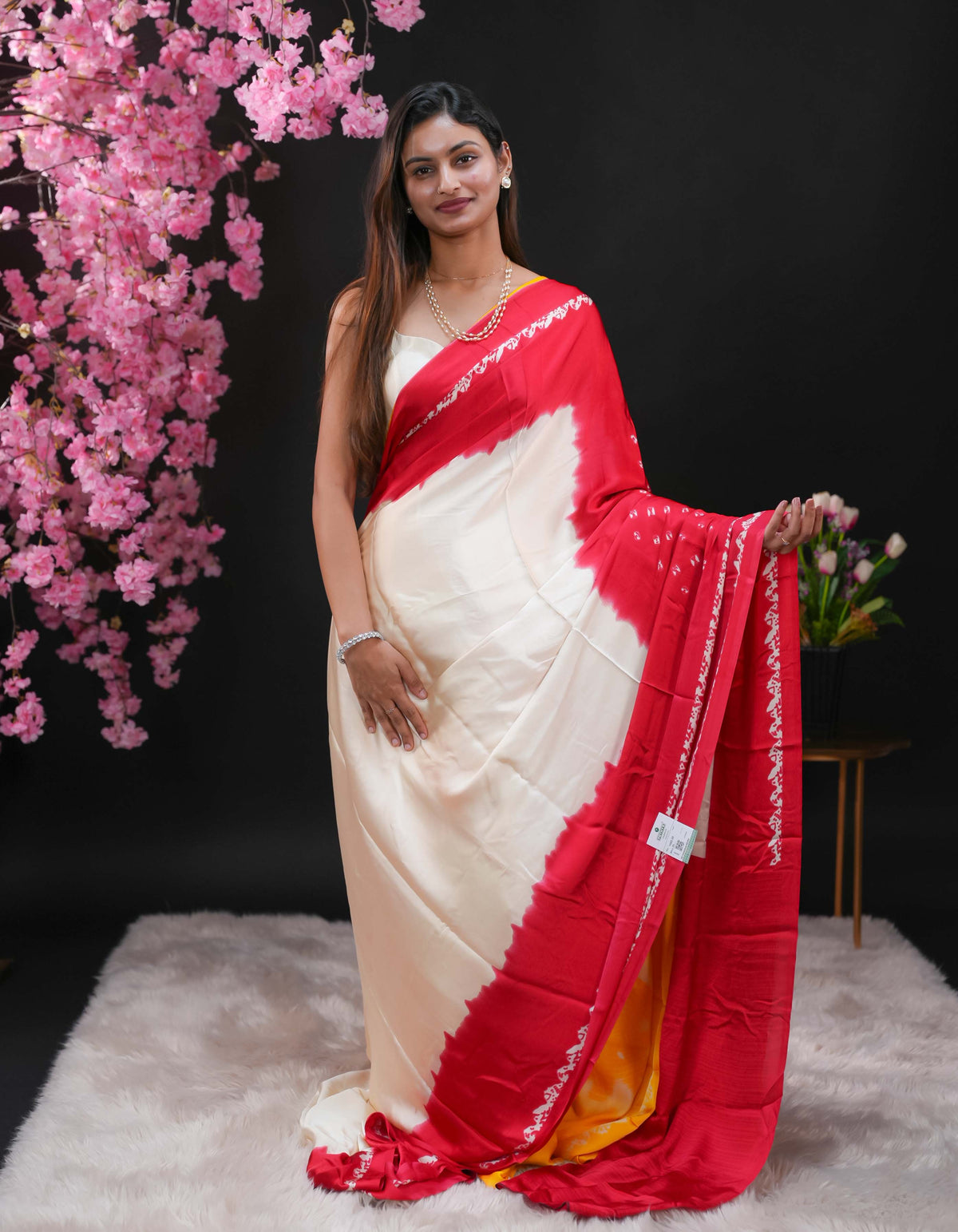 Deep Red coloured modal satin saree featuring a plain cream coloured body, complemented by a double shaded border and a Bandhani designed pallu with jhumka tassels. Paired with a matching contrast coloured blouse piece.