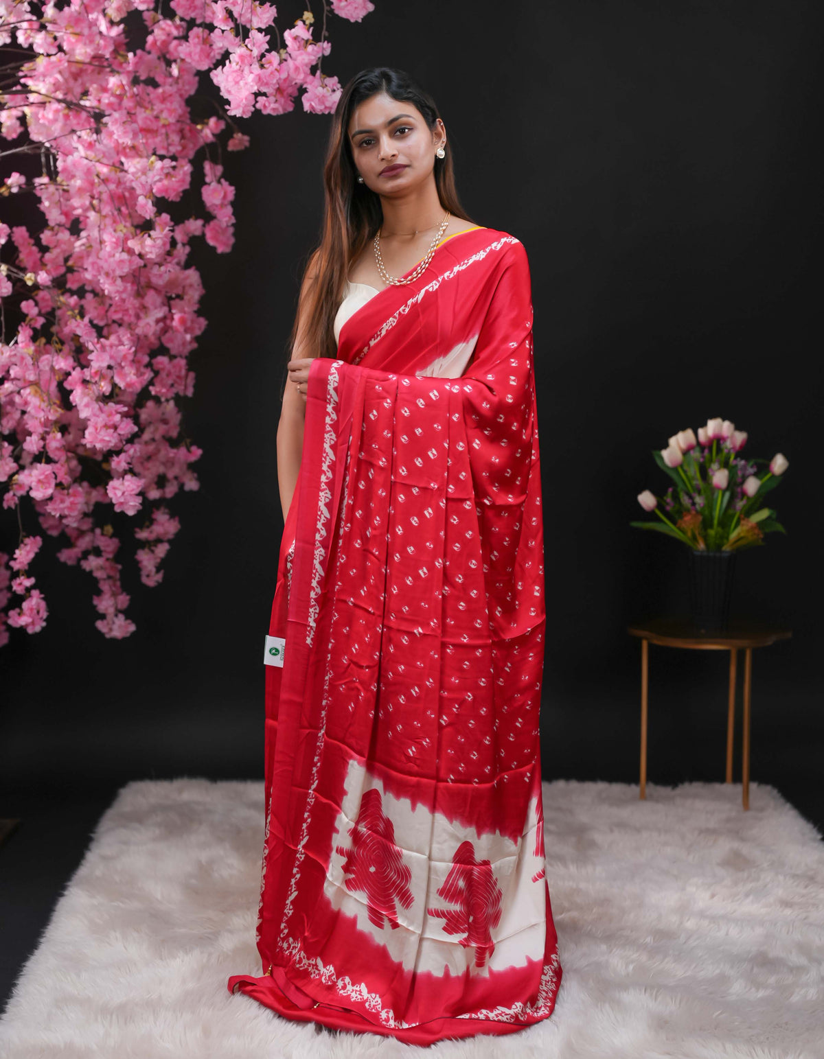 Deep Red coloured modal satin saree featuring a plain cream coloured body, complemented by a double shaded border and a Bandhani designed pallu with jhumka tassels. Paired with a matching contrast coloured blouse piece.