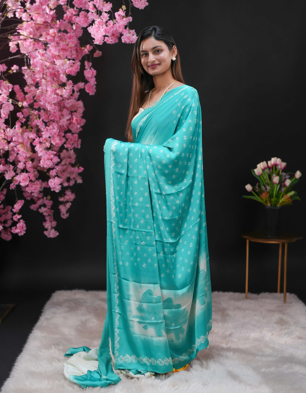 Teal coloured modal satin saree featuring a plain cream coloured body, complemented by a double shaded border and a Bandhani designed pallu with jhumka tassels. Paired with a matching contrast coloured blouse piece.